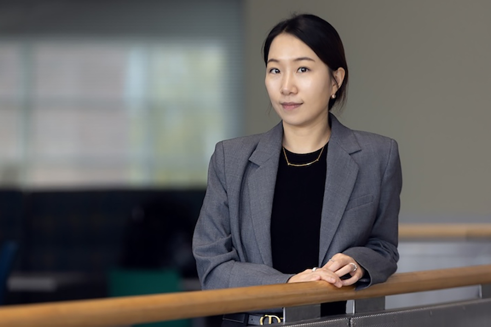 Person wearing a gray blazer and black top standing indoors near a railing in an office or academic building.