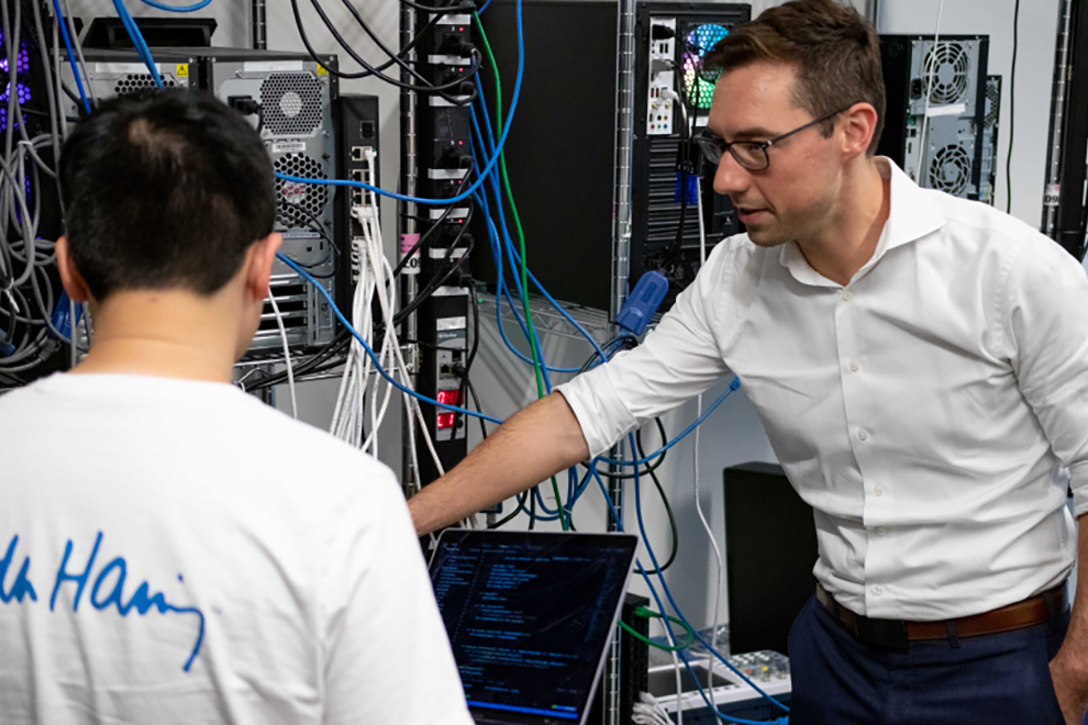 Two people working in a server room surrounded by network cables and computer hardware while reviewing code on a laptop.