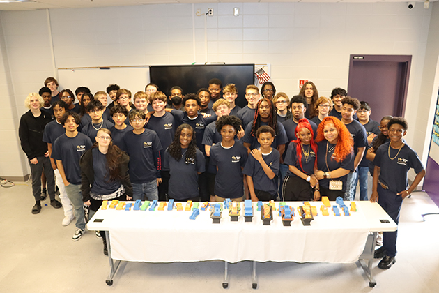 Large group of students posing for a photo behind a table displaying multiple colorful 3D-printed parts arranged in rows.