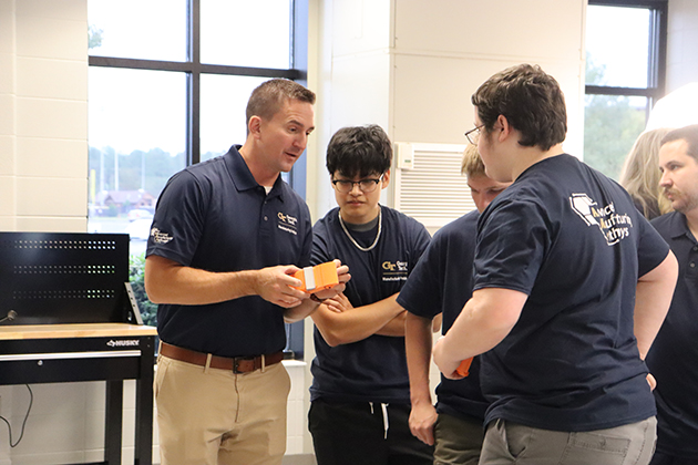 Instructor demonstrating a small orange object to a group of students wearing matching navy shirts in a classroom with large windows.
