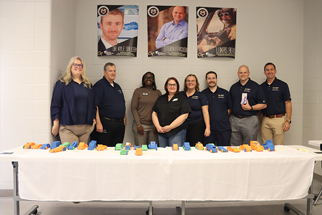 Group of instructors standing behind a table covered with colorful 3D-printed objects, with posters of professionals displayed on the wall behind them.