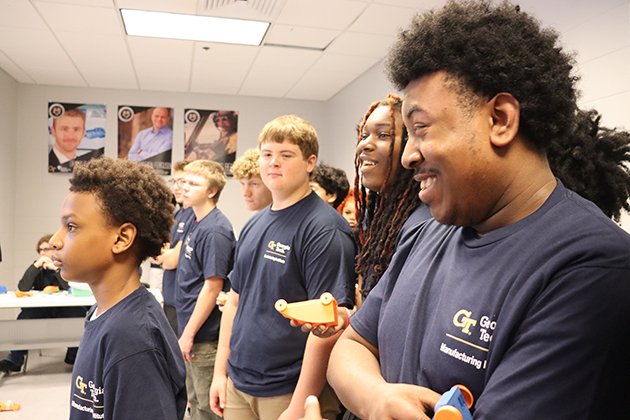 Group of students in navy shirts standing in a classroom, each holding small orange and blue 3D-printed objects.