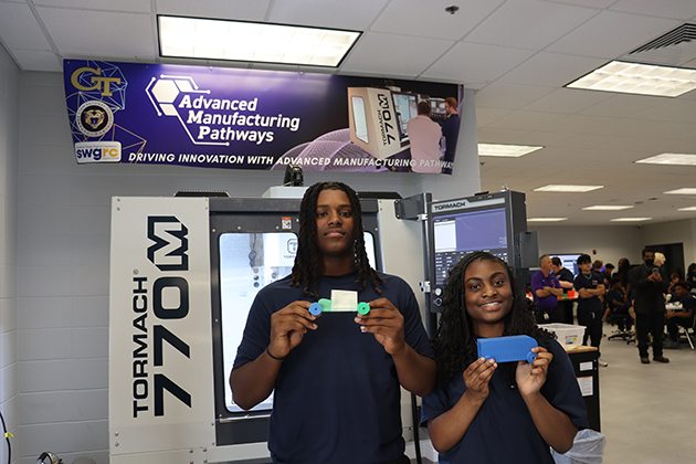 Two students holding colorful 3D-printed parts in front of a Tormach 770M machine inside an advanced manufacturing lab with a banner reading “Advanced Manufacturing Pathways.