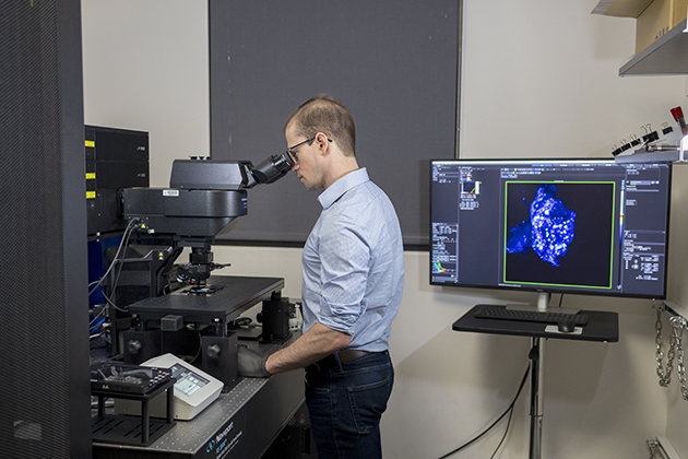 A person stands at a laboratory workstation operating a microscope, with a nearby computer monitor displaying a magnified blue scientific image.