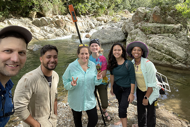 group of six people standing in front of a rolling river and bank with stones