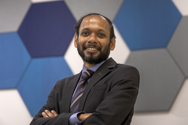 Man wearing a black suit posing with arms crossed while standing in front of multiple blue and gray hexagon tiles