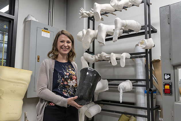 A woman holding a black prosthetic socket in a research lab, with shelves behind her displaying multiple white plaster limb molds.
