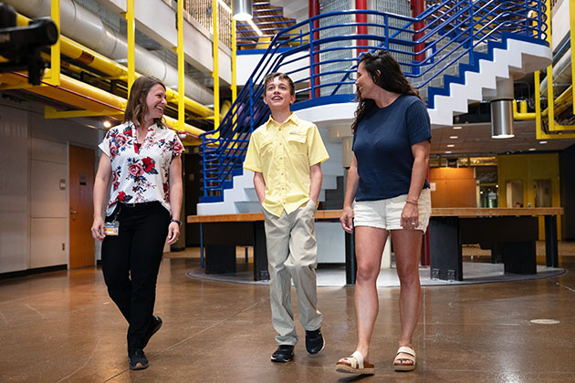 Three people walking together inside a modern building with colorful yellow and blue railings and an industrial-style interior.