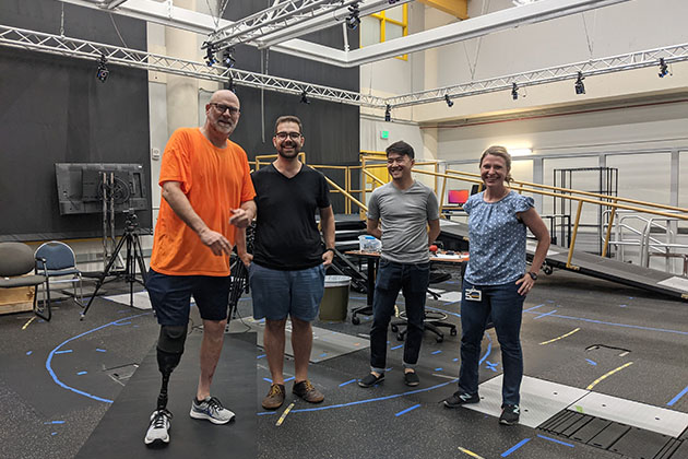 Four people standing in a lab-like environment, with one person wearing an orange shirt and a prosthetic leg, surrounded by equipment and marked floor areas.