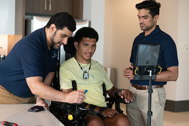 Two individuals assist a wheelchair user using an assistive device while a tablet mounted on a stand is positioned nearby in an indoor setting.