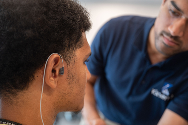 Close-up of a person wearing an earpiece device while another individual positions or adjusts equipment during an indoor session.