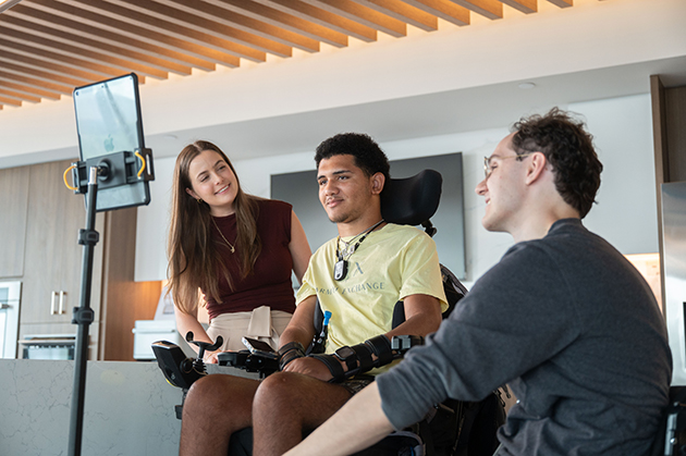 Individual using a motorized wheelchair indoors with two others nearby and a mounted tablet device positioned in front for interaction or communication.
