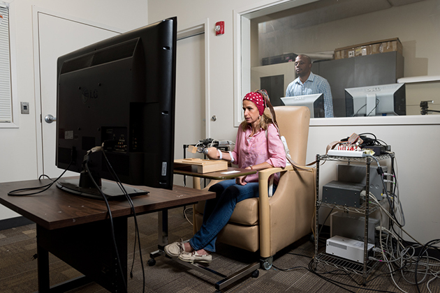 Person seated in a research lab chair using hand-operated devices connected to wires, facing a large monitor, with additional equipment and computers visible in the background through a window.