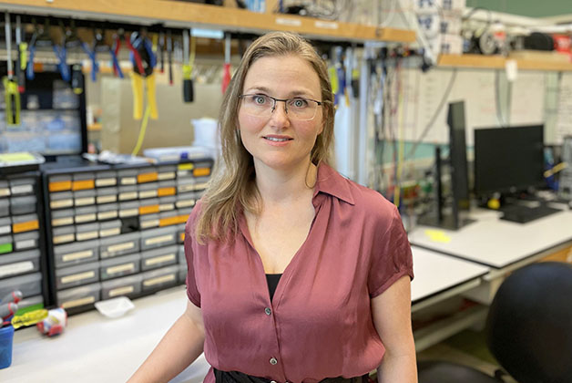 Person standing in an electronics lab with organized shelves of components and tools behind them, and a workbench with equipment in the foreground.