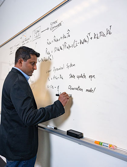 Person writing mathematical equations and system models on a large whiteboard in an office or classroom setting.