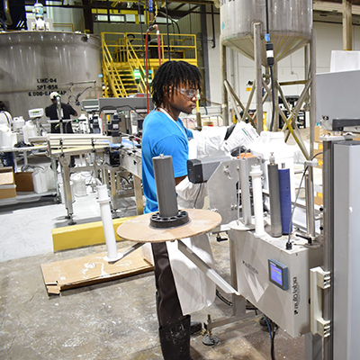 young male researcher standing in the middle of an industrial manufacturing facility surrounded by shiny metal equipment