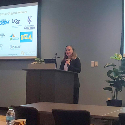 female wearing a suit standing at a podium giving a presentation with two projection screens behond her