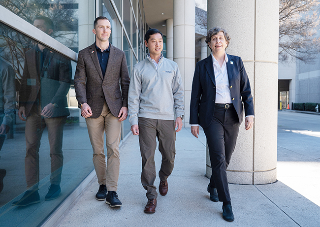 Three people walking together outside a modern building with large glass windows and concrete columns.