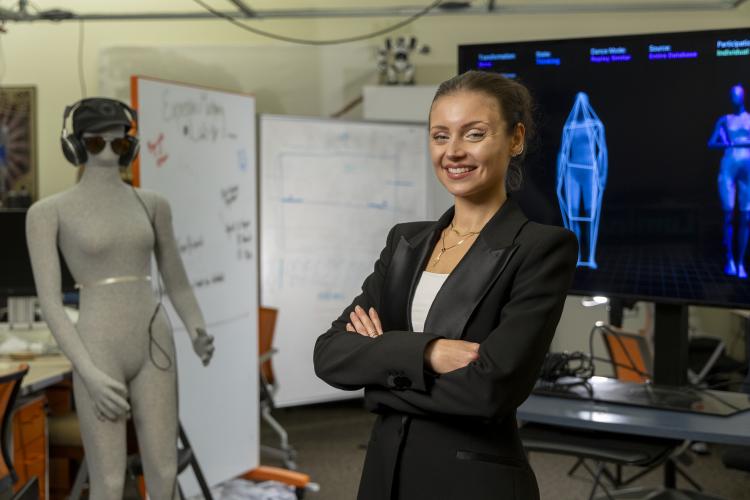 female with her hair pulled up and wearing a black suit jacket standing in front of two screen monitors showing blue outlines of human forms