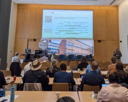 A view inside the Scholars Event Theater of a session of the Sustainability Showcase. A man speaks to a crowd while presenting slides on a large projection screen.