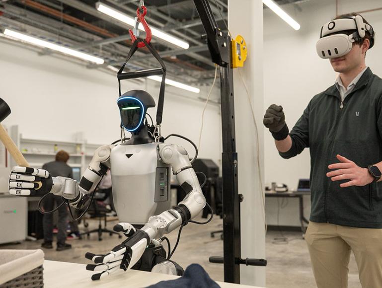 Person wearing a virtual reality headset controlling a humanoid robot equipped with tools in a laboratory setting.