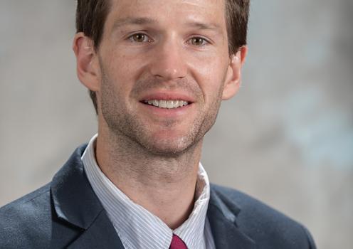 A man with brown hair and a short beard smiles for a portrait while wearing a dark blue suit and red tie.