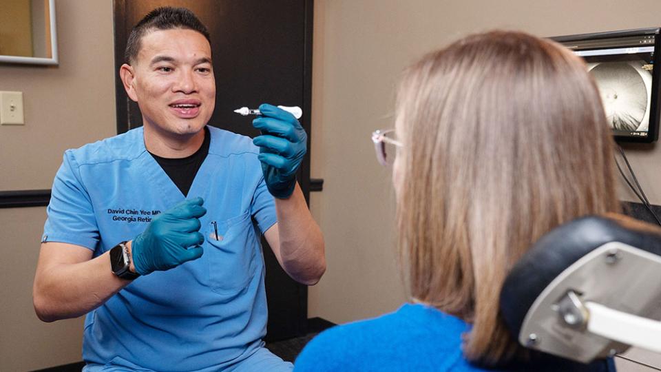 Person in blue medical scrubs demonstrates a small device to a seated patient in a clinical exam room with medical equipment visible in the background.