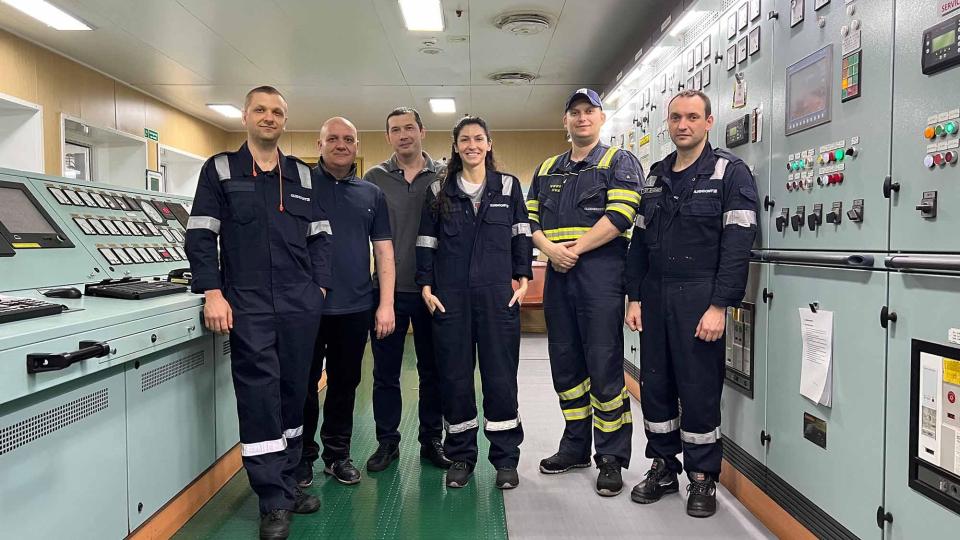 Six people in dark blue boiler suits standing in the control room of a ship