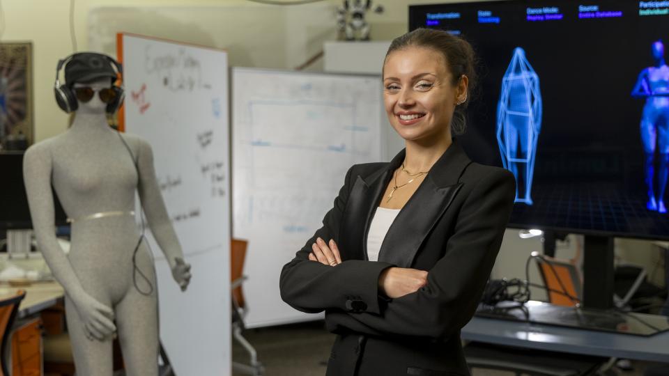 female with her hair pulled up and wearing a black suit jacket standing in front of two screen monitors showing blue outlines of human forms