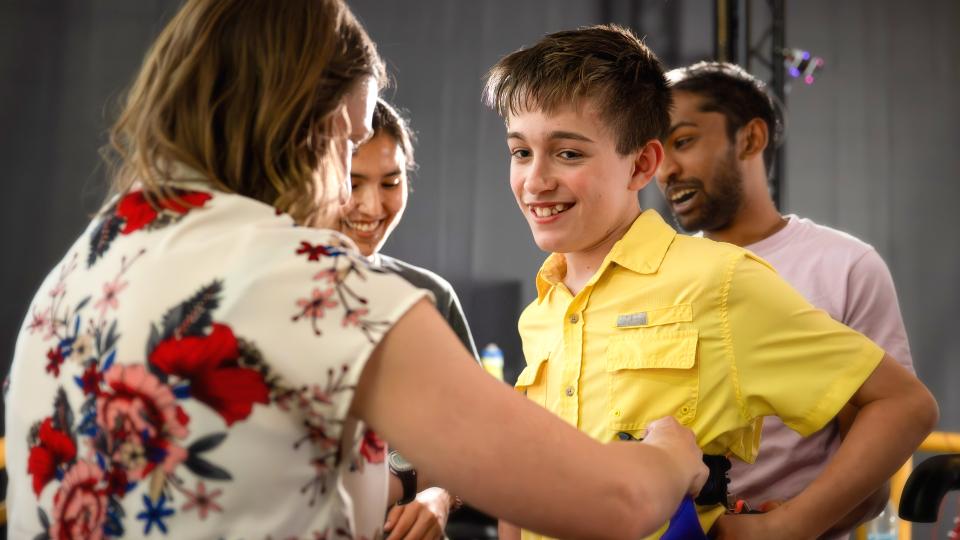 Person wearing a floral-patterned shirt interacting with a group of people indoors; one individual is dressed in a bright yellow button-up shirt.