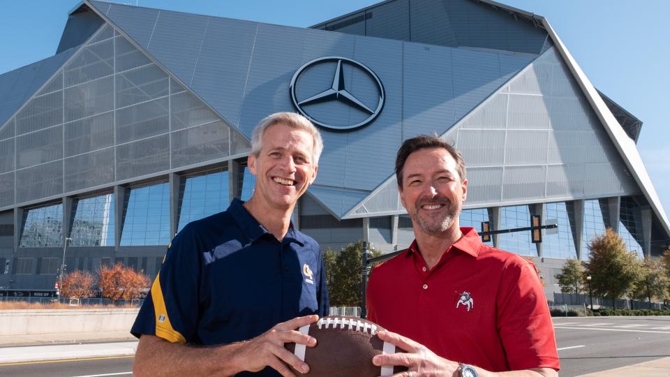 A tall white man wearing a blue GT-branded polo standing next to a slightly shorter man wearing a UGA-branded red polo. They're smiling and both holding a football.