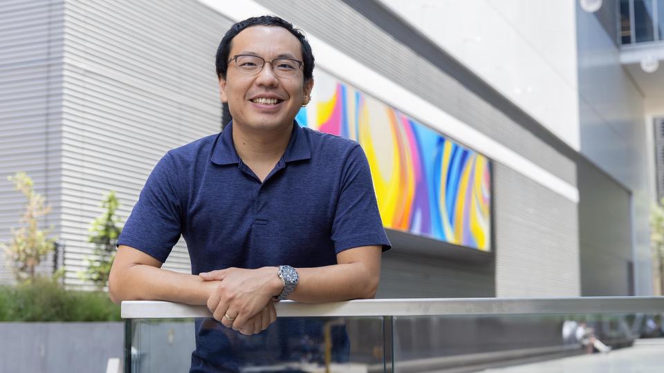 A man standing outside in a building breezeway. He is wearing glasses, a blue polo and is smiling.