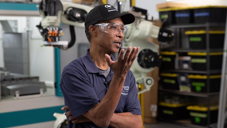 A man wearing a cap, safety goggles, and a navy shirt speaks in an industrial setting.