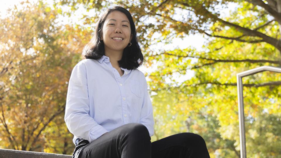 Person sitting outdoors on stone steps wearing a light blue button-up shirt and black pants, with autumn trees and sunlight in the background.