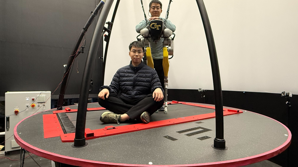 Two individuals on a large circular motion platform in a research lab, with one person seated cross‑legged on the platform and another suspended in a harness wearing a Georgia Tech exoskeleton system.