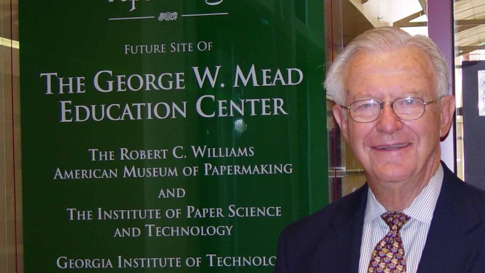 Older man in suit, smiling, stands next to a green sign with white text. 