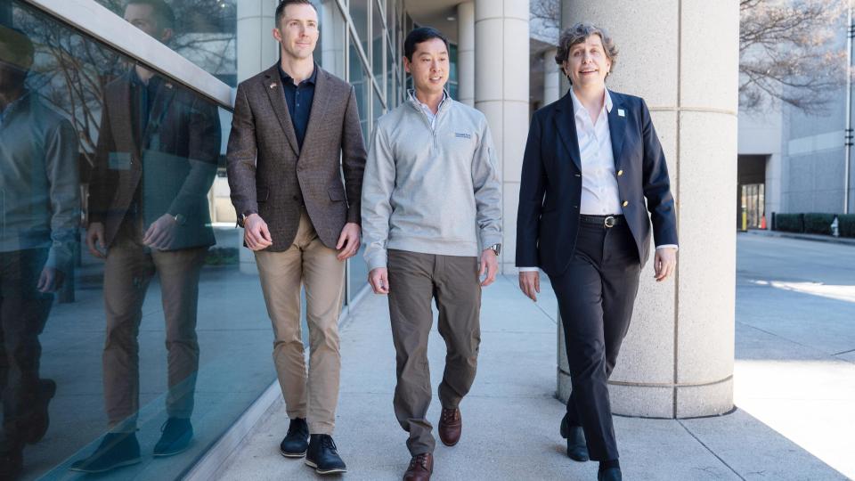 Three people walking together outside a modern building with large glass windows and concrete columns.