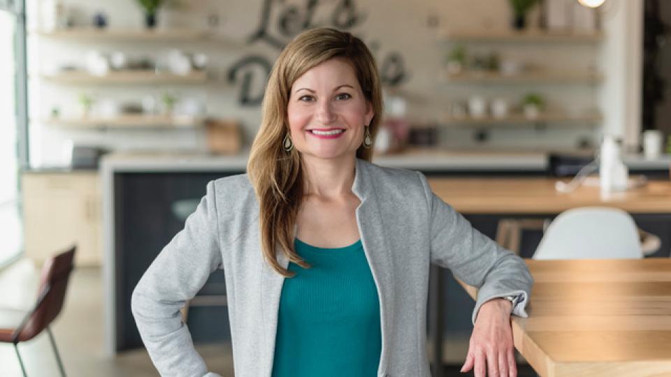 Alison Sizer in a blazer standing in a modern workspace with wooden tables, open shelving, and natural light.