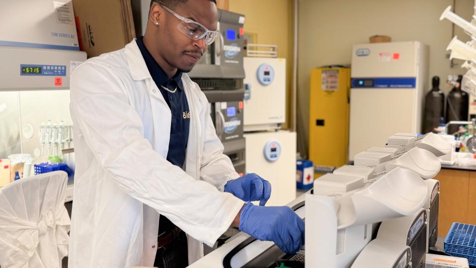 A man in a lab coat wearing safety goggles and gloves puts samples into a machine in a scientific lab