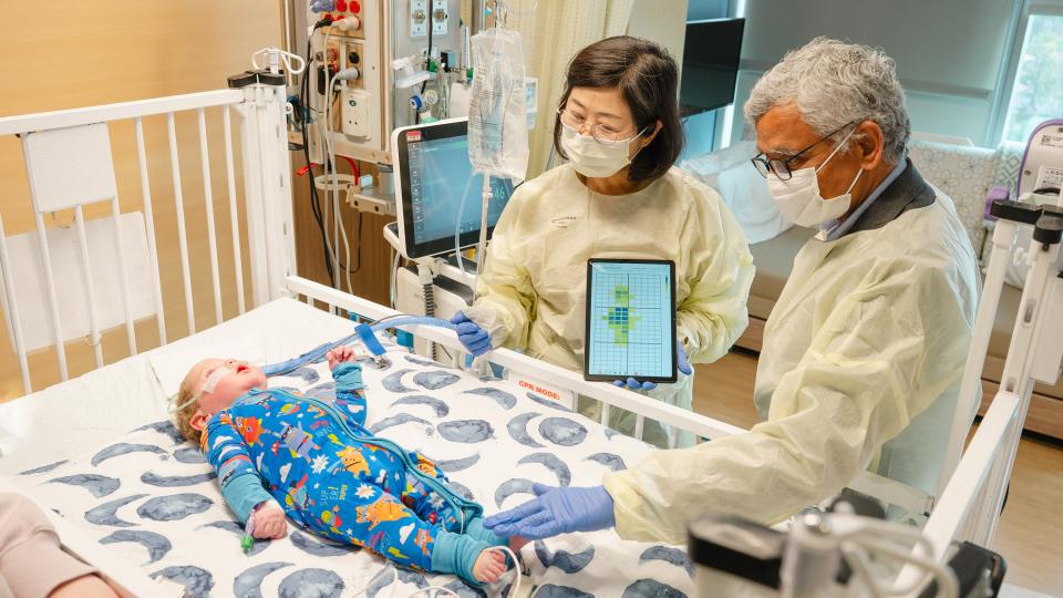 Two adults wearing protective gowns and gloves stand beside a hospital crib, using a tablet device while examining an infant lying on the mattress as medical equipment and monitors surround the crib.