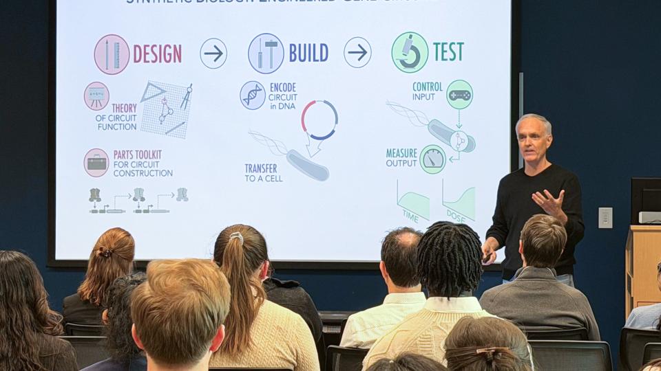 A presenter stands at the front of a lecture room speaking to a seated audience while a projected slide titled “Synthetic Biology: Engineered Gene Circuits” illustrates the design–build–test cycle with diagrams and icons explaining gene circuit construction and testing.