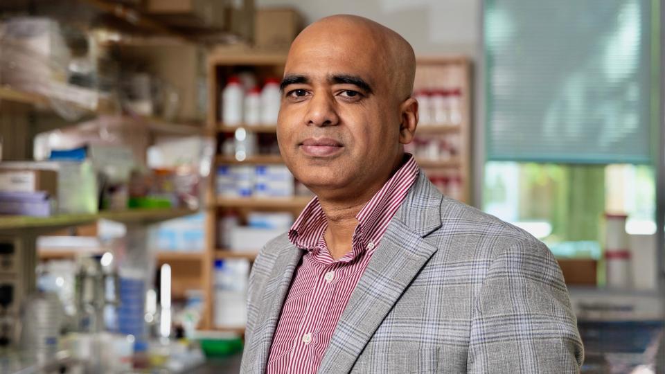 Ankur Singh, a man in a gray suit jacket with a dark pink button-up shirt stands in front of a work bench in a lab.