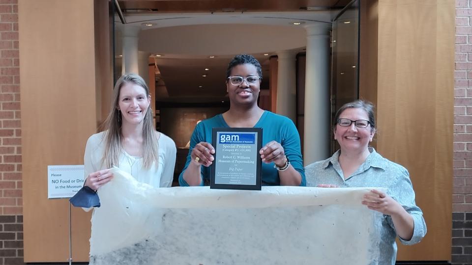 Three museum staff stand in front of the museum doors holding a very large sheet of paper and a plaque. 