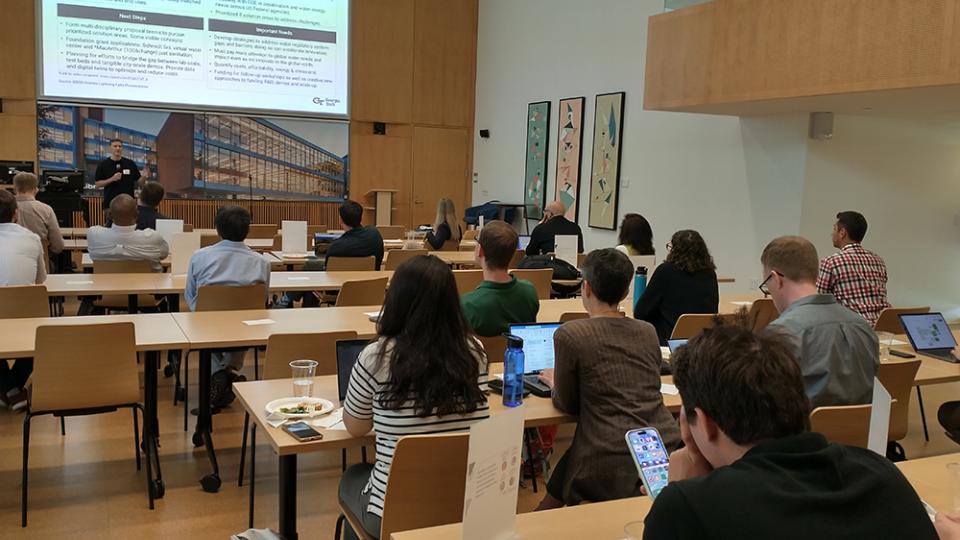 View of a man presenting to a crowd of onlookers in a large meeting room.
