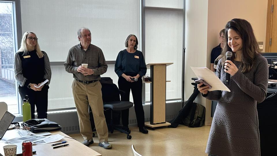 "A group of people stand near large windows in a bright indoor space, with one person holding a microphone and reading from a sheet of paper. Three individuals stand together near a lectern, some holding notes or drinks. In the foreground, a table holds laptops, papers, and a drink cup. The room has light-colored floors and tall window shades that diffuse the natural light."