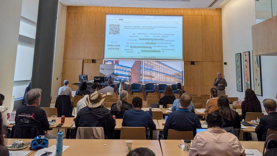A view inside the Scholars Event Theater of a session of the Sustainability Showcase. A man speaks to a crowd while presenting slides on a large projection screen.