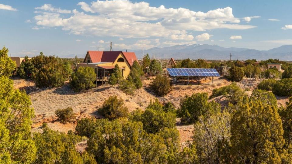 A rural residence with solar panels installed outdoors, set among desert vegetation with mountains in the distance.