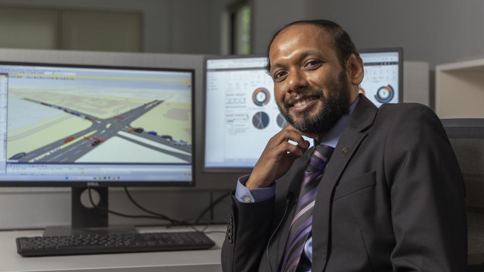 Angshuman Guin (a male professor wearing a black suit) sits at a desk in front of two monitors displaying data