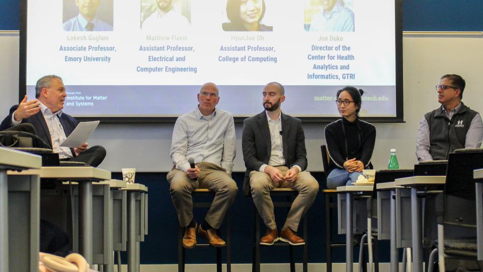 A panel of five speakers sits on tall stools at the front of a classroom, participating in a moderated discussion. The moderator on the left holds papers while addressing the group. A large presentation slide behind the panel displays names and academic titles. Audience members are partially visible in the foreground, and tables, chairs, and a water bottle are arranged throughout the room.