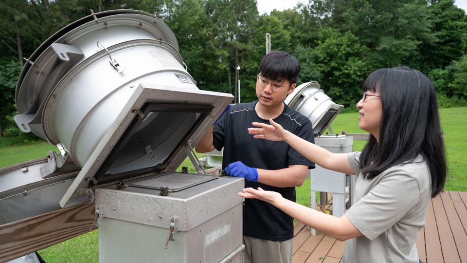A male and female researcher working with a metal piece of equipment outdoors with trees and grass in the background